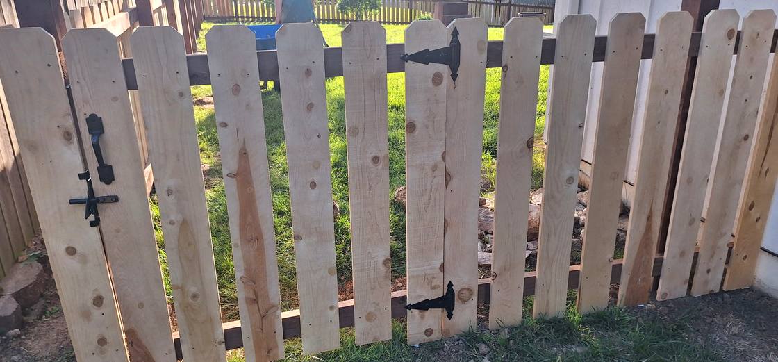 A cedar picket gate hung on an ornate black antique style strap hinge, slim against the boards rather than a bulky box hinge.